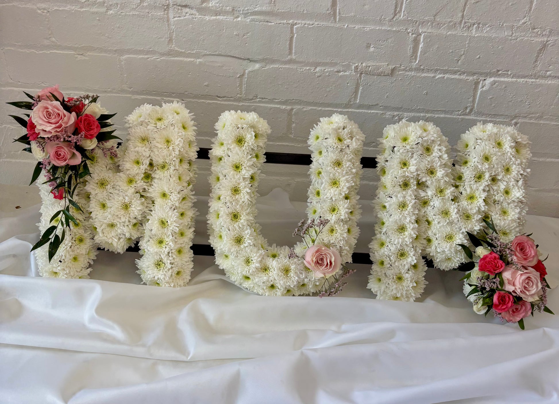Large decorative letters spelling “MUM” filled with white chrysanthemums, displayed on a white draped surface against a white brick wall, with pink rose floral arrangements attached to the left and right sides.