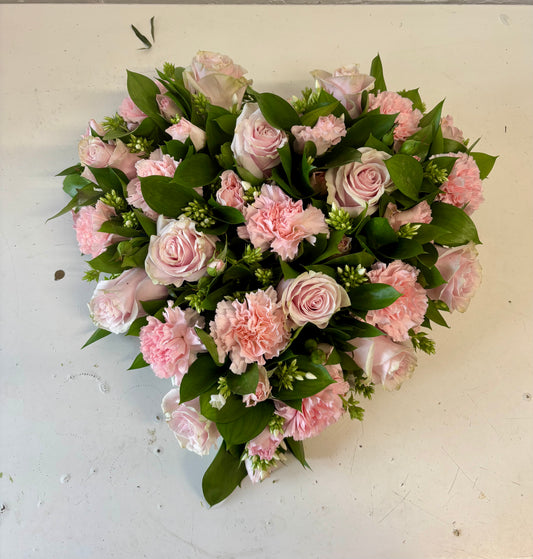 A heart-shaped floral arrangement featuring light pink roses, pink carnations, and green foliage on a light off-white surface.