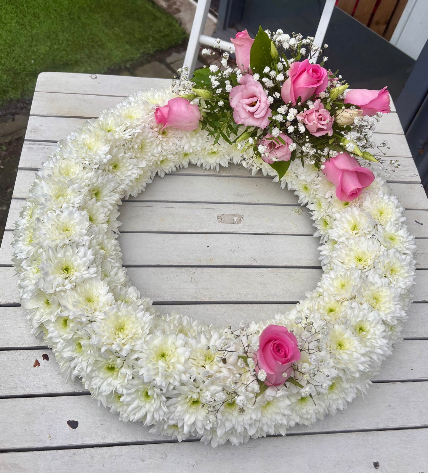 A circular floral wreath made of white chrysanthemums with pink roses and baby's breath, displayed on a white wooden table outdoors.