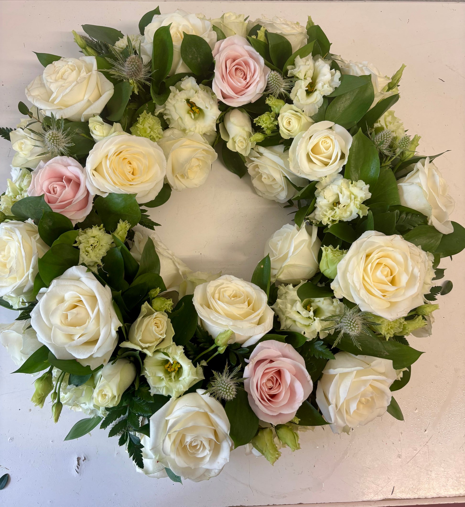 A circular floral wreath featuring white and pale pink roses, green foliage, and small light green flowers, arranged on a plain off-white surface.