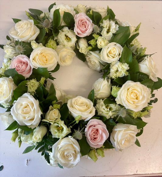 A circular floral wreath featuring white and pale pink roses, green foliage, and small light green flowers, arranged on a plain off-white surface.